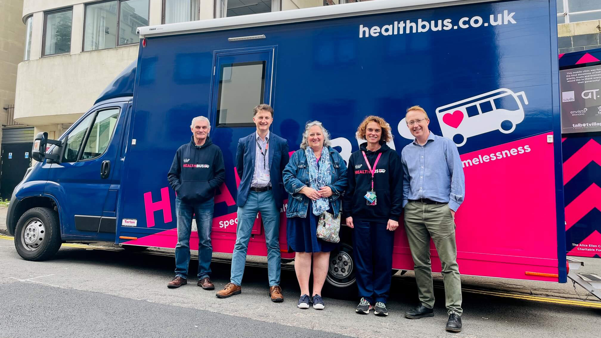 Three men and two women stand smiling in front of a blue van labelled Healthbus