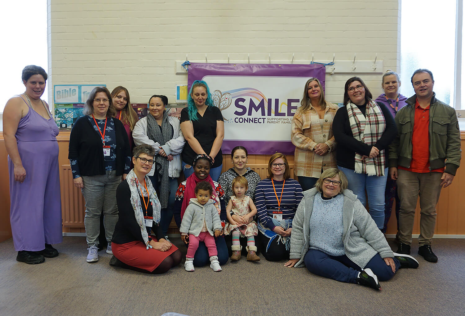 Group of women and a man standing & sitting, looking at the camera smiling. Sign in the background reads "SMILE"