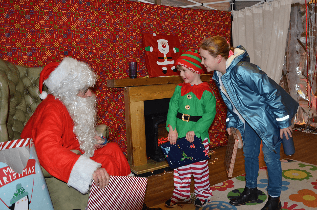 Santa is sitting in an armchair holding a present, a young boy in an elfs costume is holding a present and looking at santa, a girl next to the boy leans in towards santa smiling
