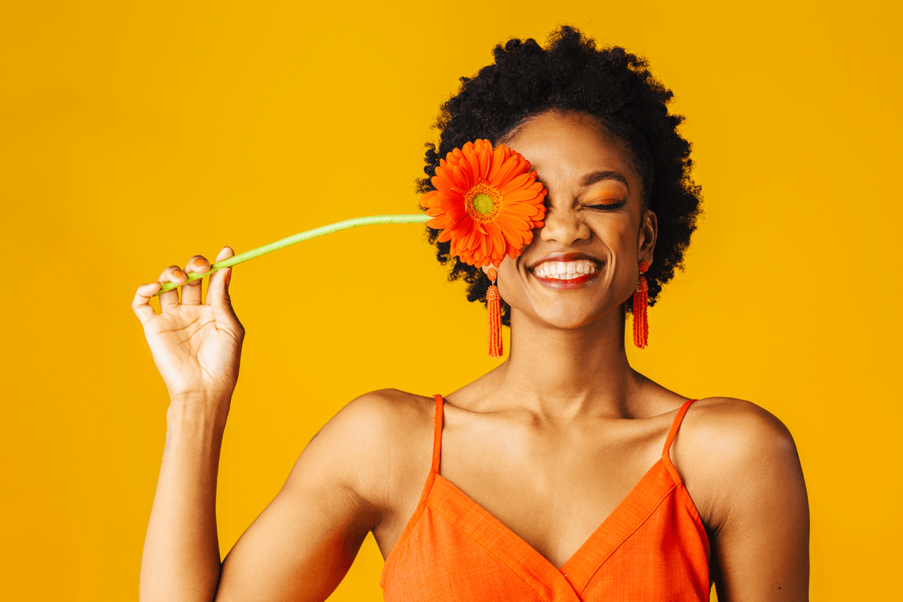Woman with her eyes shut, holding an orange flower over her eye and smiling, standing against an orange background