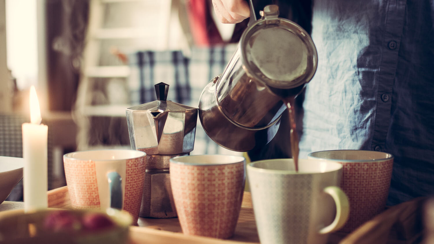 Someone pouring coffee into four mugs on a wooden tray, with a stovetop espresso maker standing next to the mugs