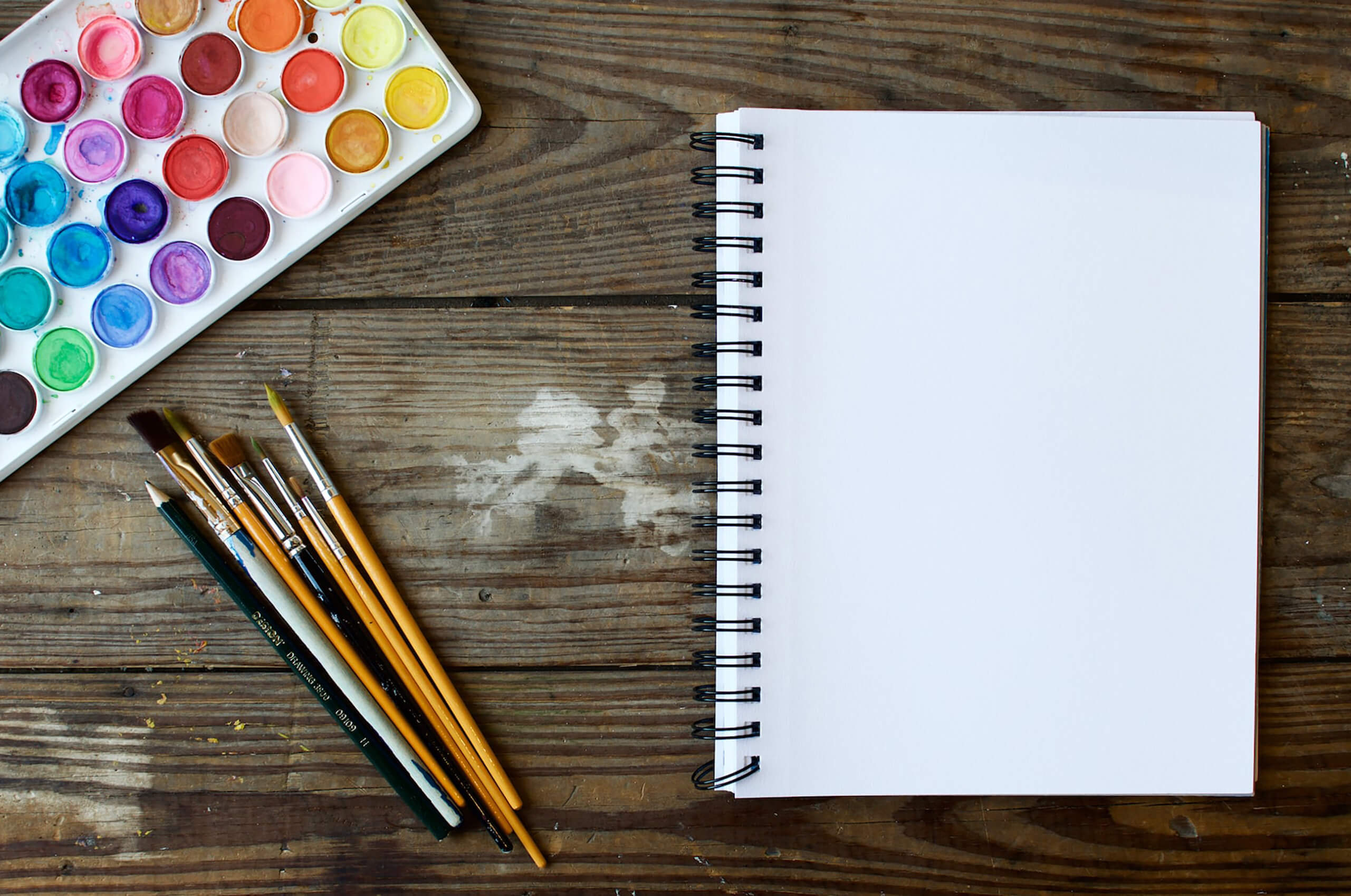 A blank sketchbook lying on a wooden table, nest to watercolour paints and paint brushes