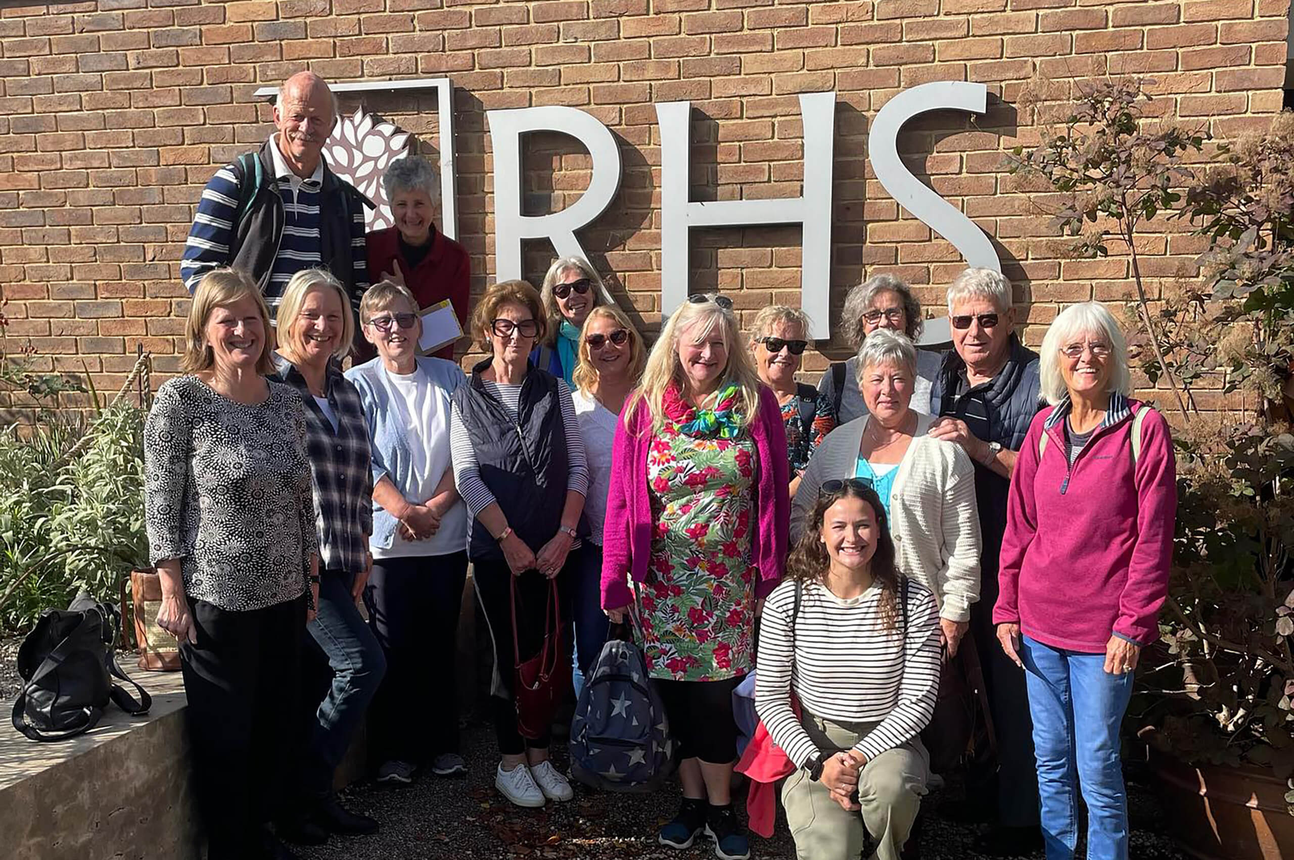 A group of women and men stand in front of a brick wall which features the logo for RHS Garden Wisley