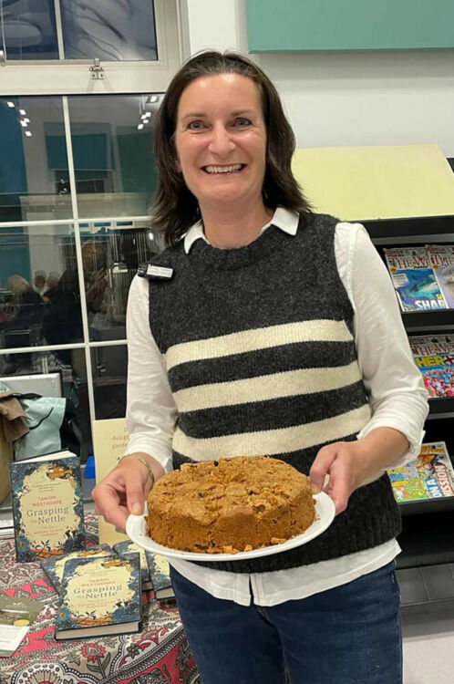 A woman stands, holding a cake and smiling at the camera