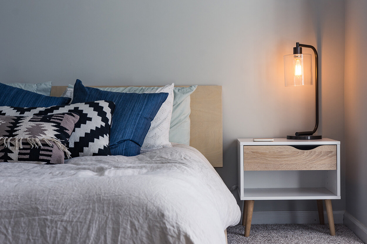 Several pillows lie on a bed, next to a side table with lamp