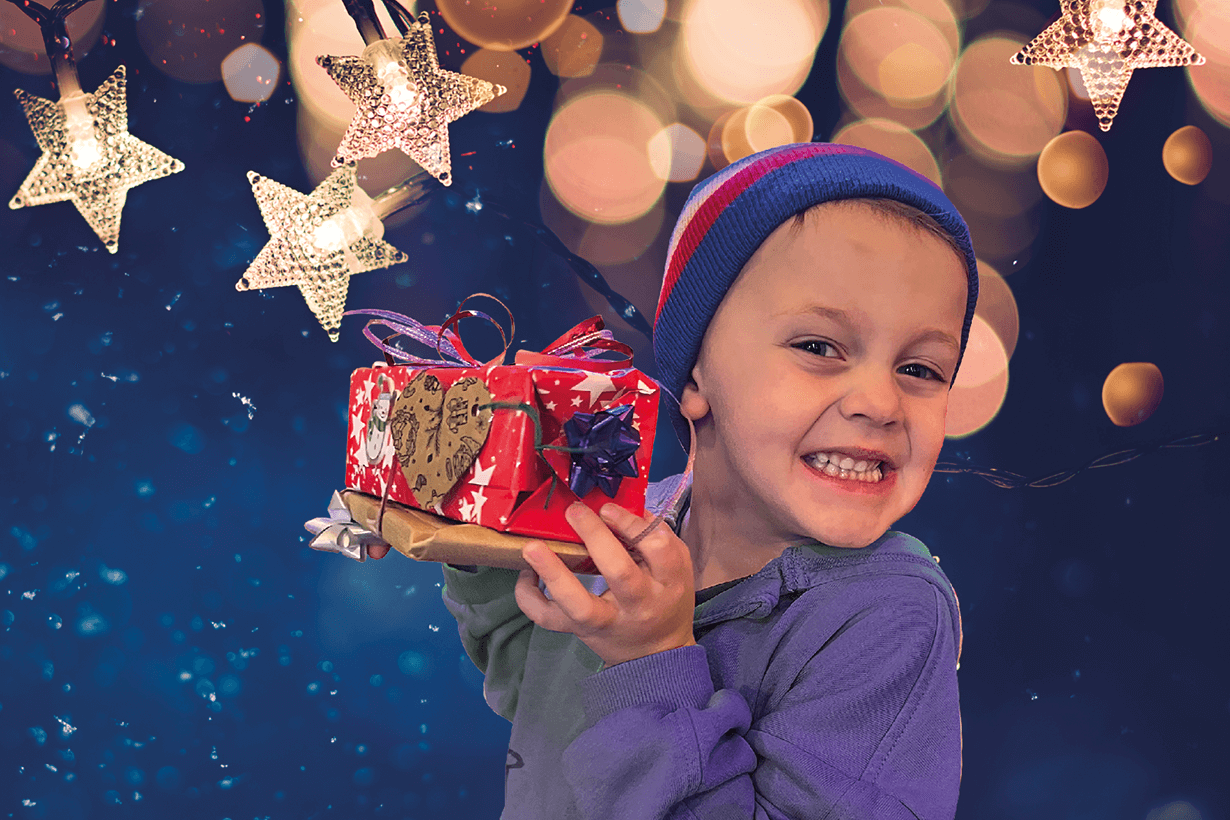 A boy wearing a hat and holding a gift wrapped Christmas present up, and grinning. The boy is standing against a blue backdrop with stars and snow