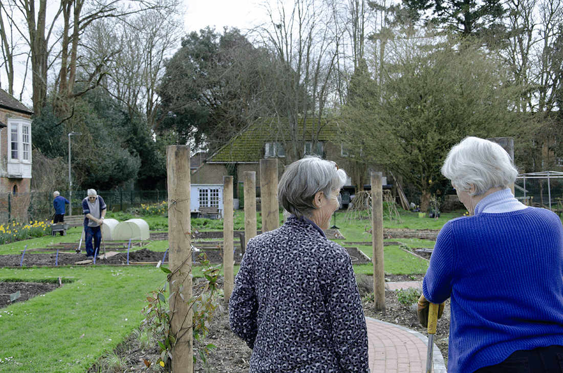 Two women stand chatting in front of a garden with two men gardening in the background
