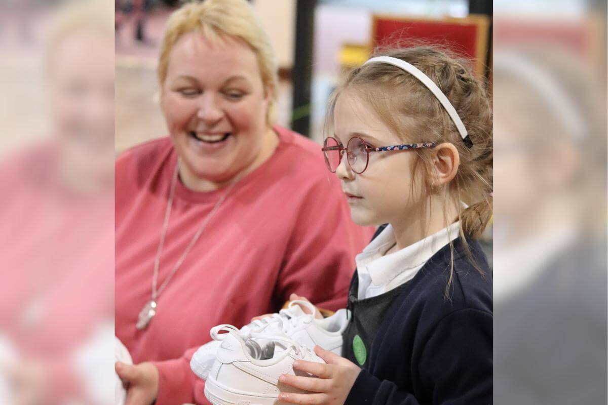 A parent smiles at a young girl holding a pair of new white trainers