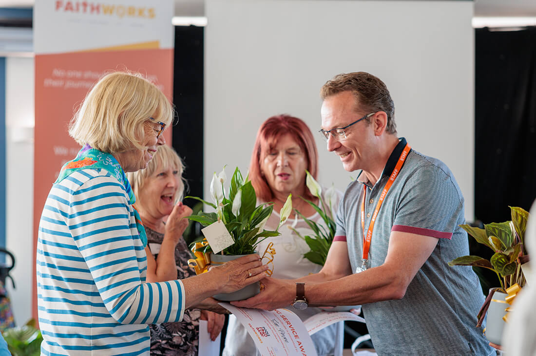 A man presents a woman with a plant, two women watch - the group look happy and are smiling