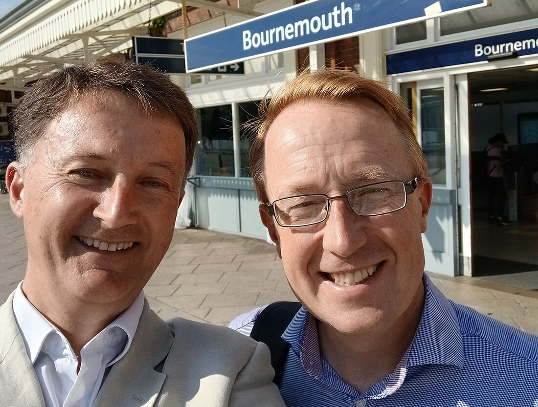 Two men stand in front of Bournemouth train station, smiling at the camera