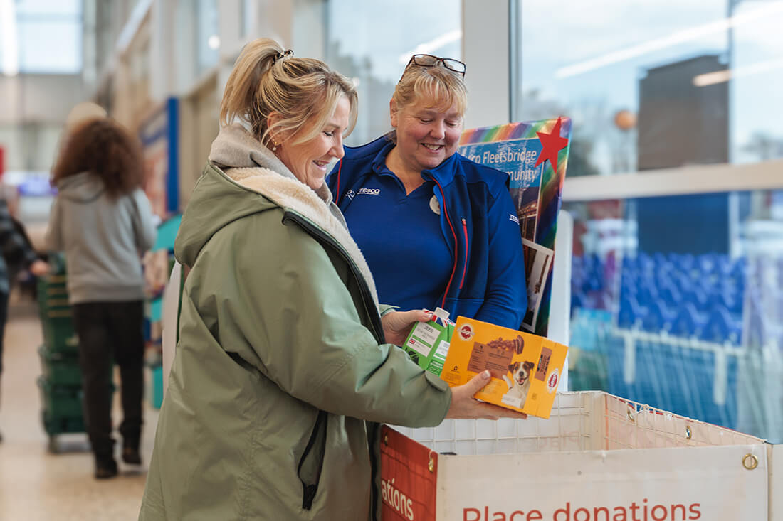 A woman smiles as she places food items into a food donation basket, while a smiling woman look son