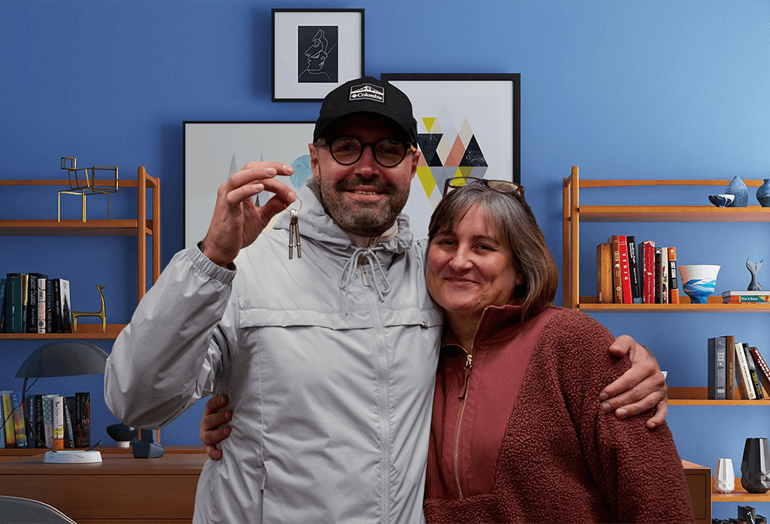 A man and a woman stand with their arms around each other, smiling at the camera. The man holds a key in his fingers.